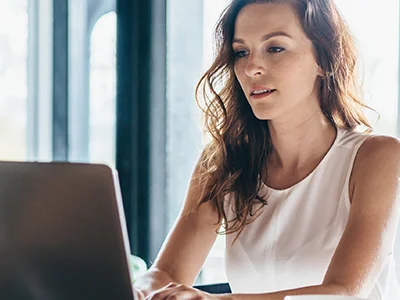 Foto einer Frau mit langen braunen Haaren und weißem ärmellosem Top sitzend vor einem Laptop.